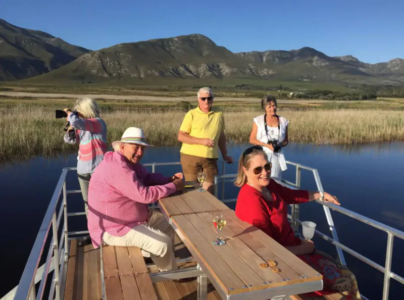 Group of smiling people on wooden deck of boat cruising Klein River toward Hermanus Estuary, mountains and wetlands backdrop.