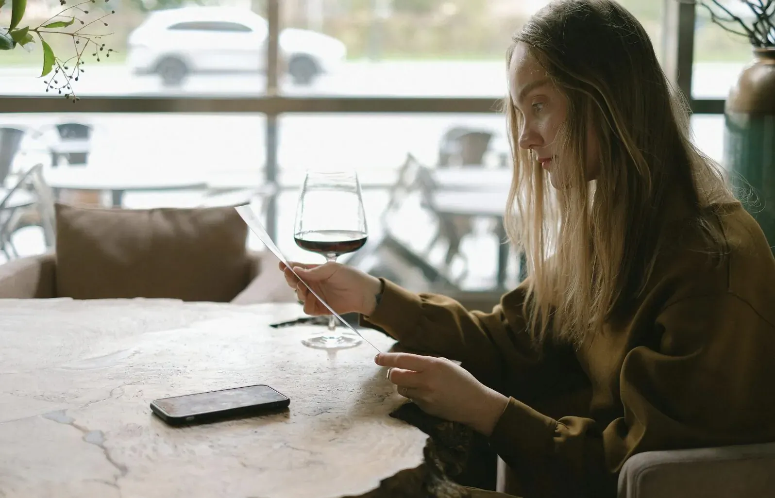 Woman in cozy cafe holding wine glass and phone, gazing thoughtfully at table with plants and window view.