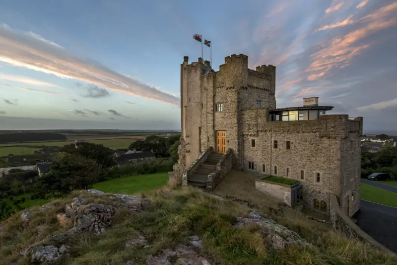 Roch Castle, a luxury stone castle hotel on grassy hilltop in West Wales at sunset with flags flying.