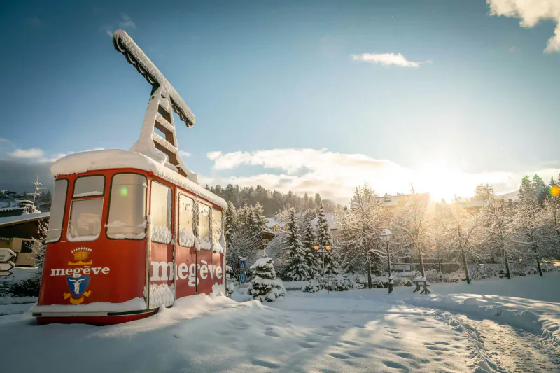 Red Megève cable car on snowy ground with mountains, pine trees, and golden sunset glow