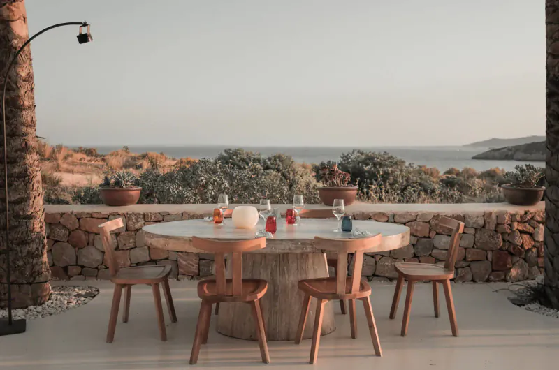 Round wooden table with colorful glass bottles and wine glasses on outdoor terrace overlooking sea at sunset, palm trees and stone walls.
