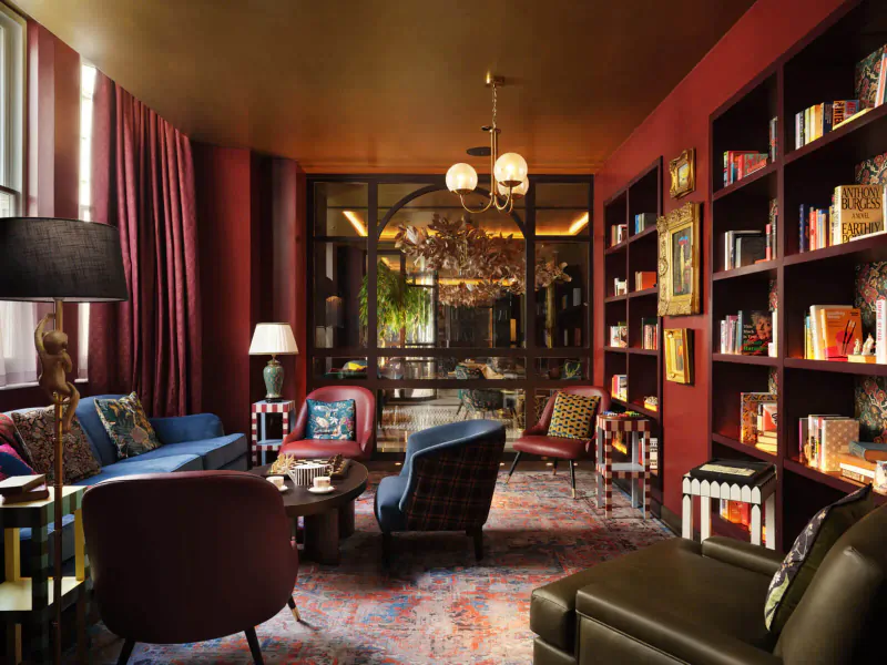 Elegant library lounge at The Other House South Kensington with red walls, bookshelves, velvet seating, chandelier, and large window.