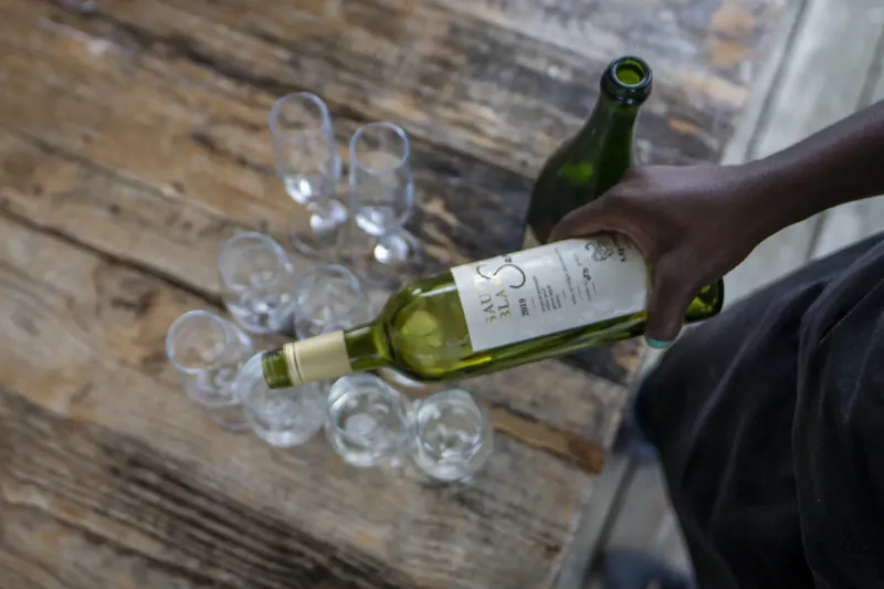 Close-up of dark hand pouring white wine from bottle labeled 'Kay & Monty' into glasses on rustic wooden table, with extra green bottle nearby.