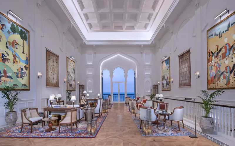 Luxurious hotel lounge with ornate Indian paintings, elegant chairs around tables, arched windows overlooking sea.