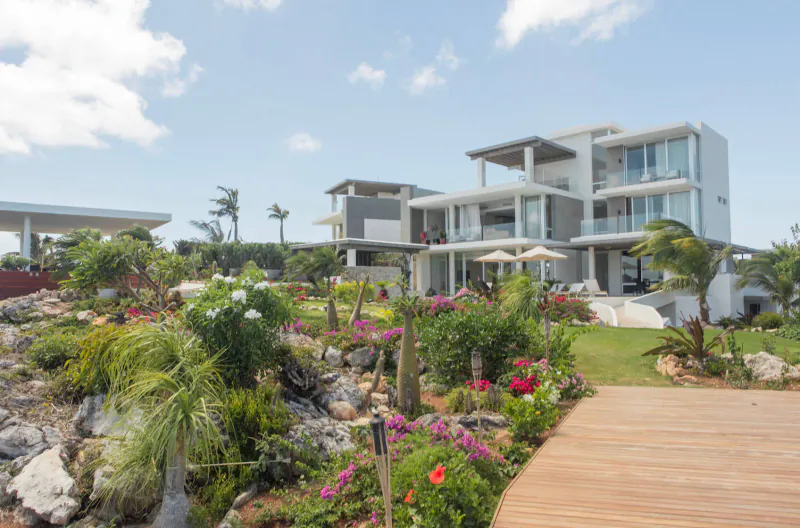 Modern white multi-level private resort house amid lush tropical gardens, palm trees, and flowers under blue sky