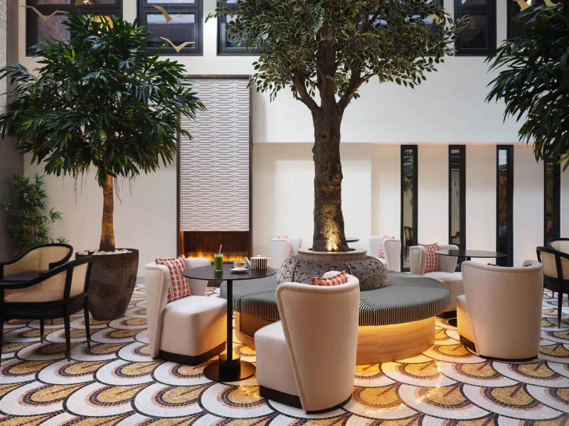 Luxurious hotel lobby with central potted tree, curved seating around glowing fire pit, potted palms, and patterned floor.