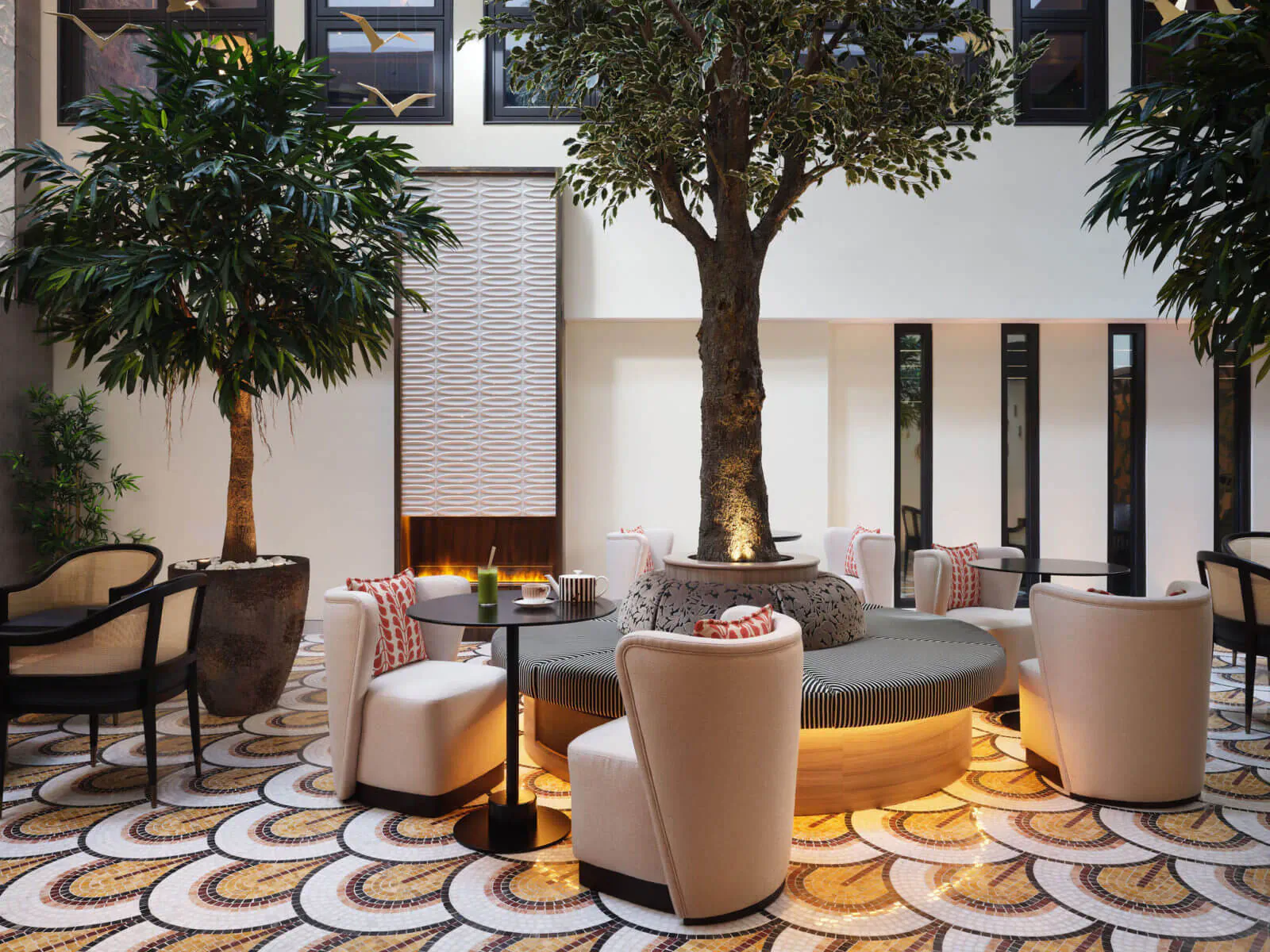 Luxurious hotel lobby with central potted tree, curved seating around glowing fire pit, potted palms, and patterned floor.