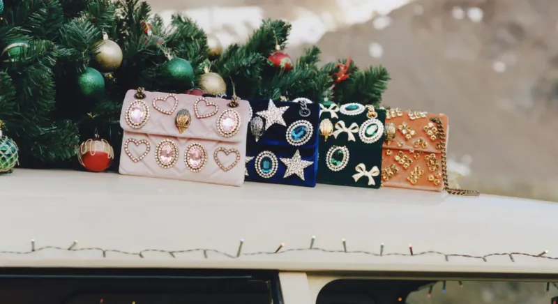 Four ornate Kurt Geiger holiday bags in pink, blue, green, gold on snowy Christmas tree branch atop white van.