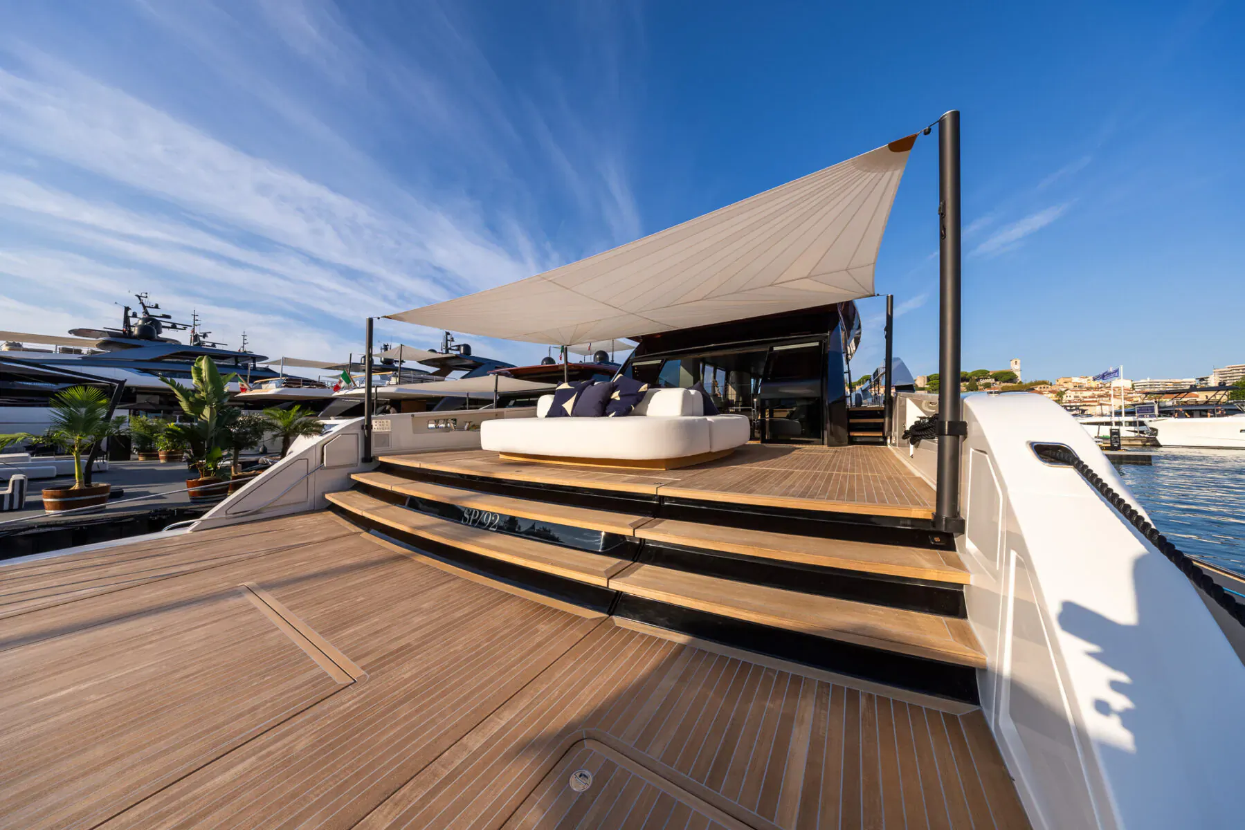 Sanlorenzo luxury yacht deck with white sunshade, cushioned seating area, wooden steps, marina backdrop under blue sky