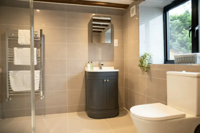Luxurious modern bathroom in The Barn with glass shower, towel rail, black vanity sink, white toilet, and window.
