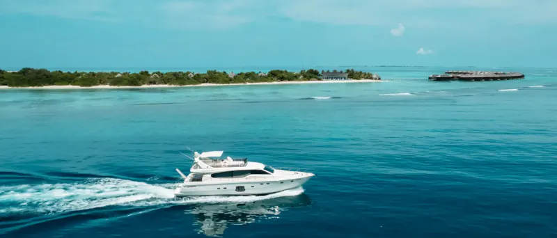 White luxury yacht speeding across turquoise ocean toward tropical island with Hideaway Majesty villas, clear blue sky.