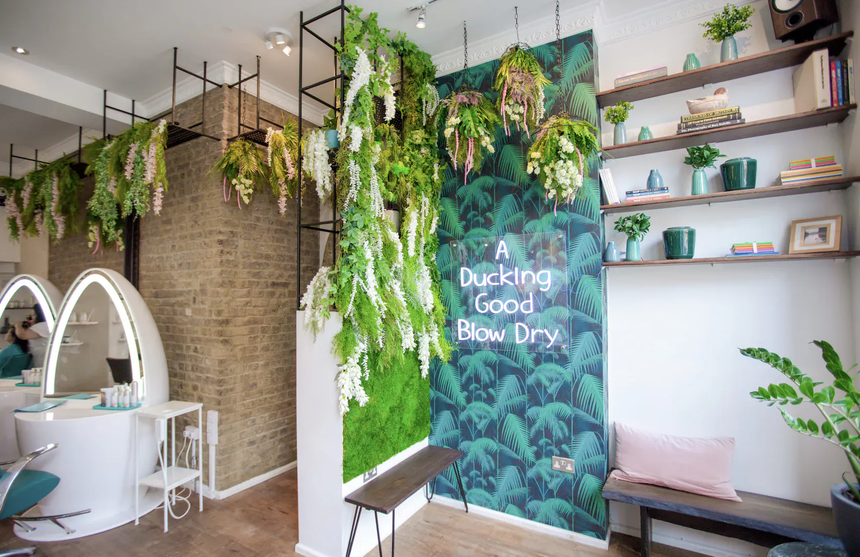 Duck & Dry salon interior with lush green plants, wisteria, moss wall featuring 'A Good Blow Dry' neon sign, mirrors, and styling stations.