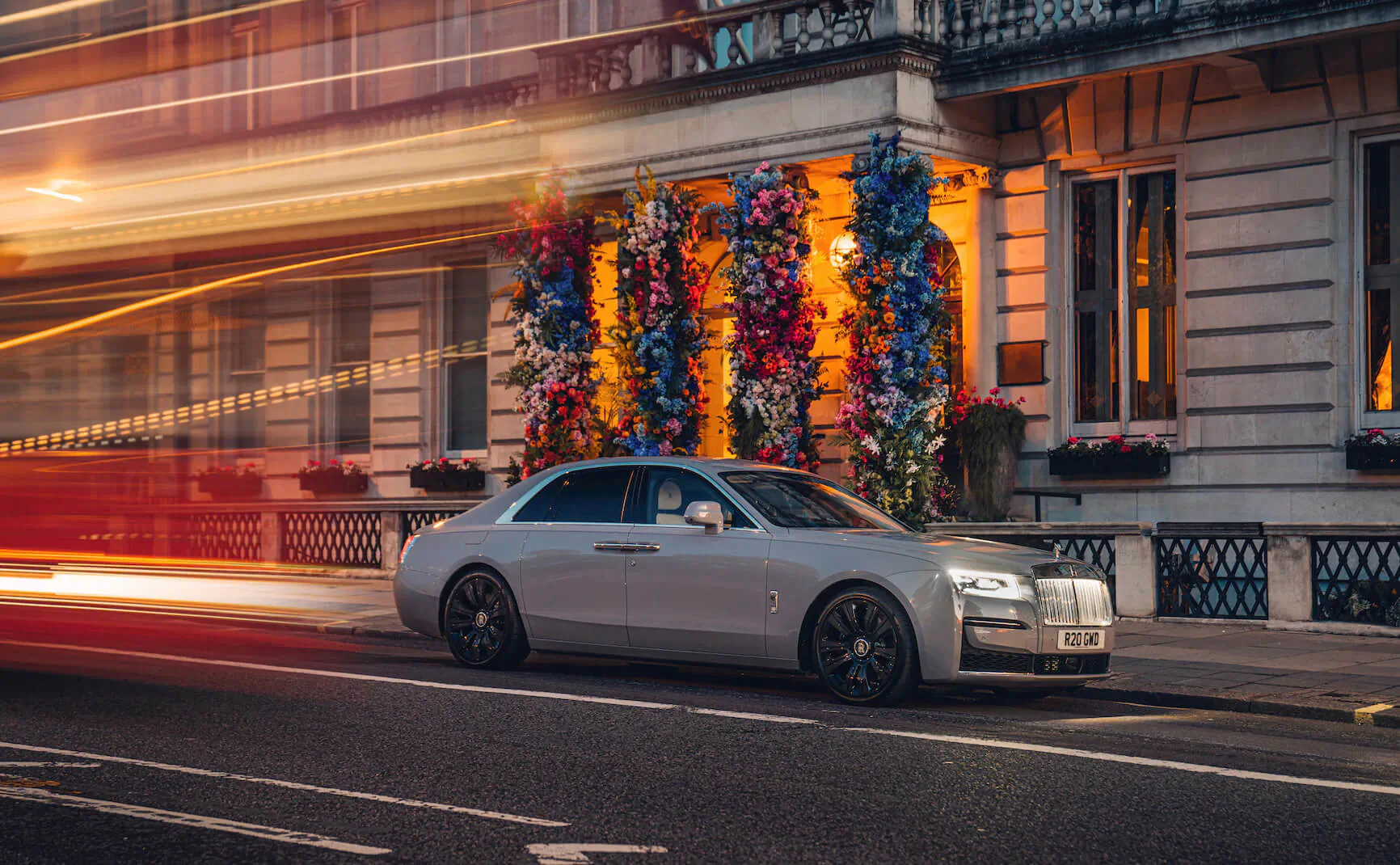 Silver Rolls-Royce sedan driving past ornate London building adorned with colorful floral garlands at night with light trails.