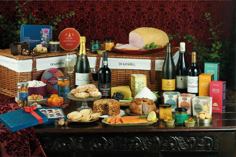 Festive Christmas hamper on wooden table with Dukeshill ham, cheeses, cakes, fruits, wine, champagne against red backdrop