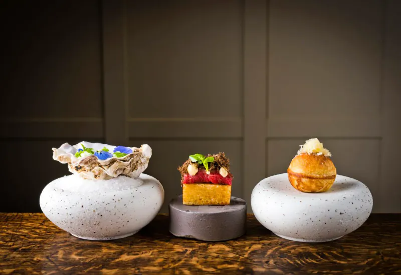 Three elegant desserts on textured white and gray stands: nest with blue leaves, beet cake with herbs, and topped pastry, on wooden table.