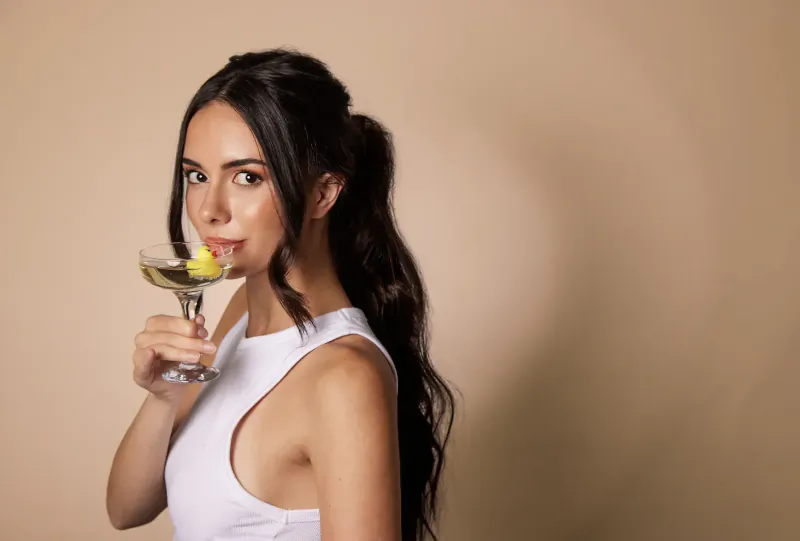 Woman with dark ponytail in white tank top holds martini with lemon twist, smiling at Galentine’s Day event
