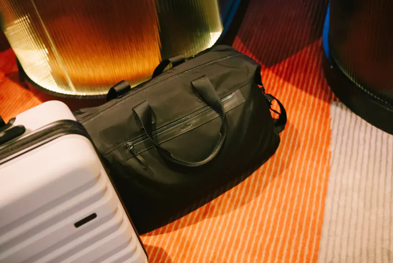 White Antler Fenchurch wheeled suitcase and black duffel bag beside illuminated columns on carpeted gallery floor