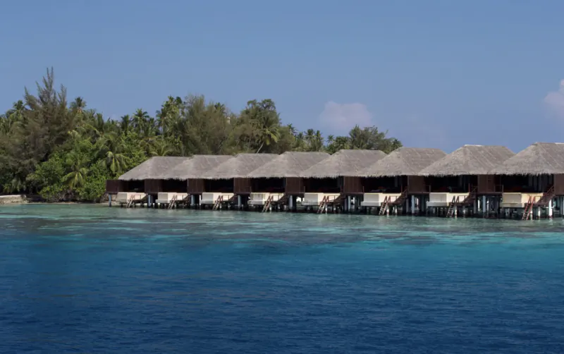 Row of thatched overwater bungalows at Coco Bodu Hithi resort, turquoise lagoon, palm trees, blue sky
