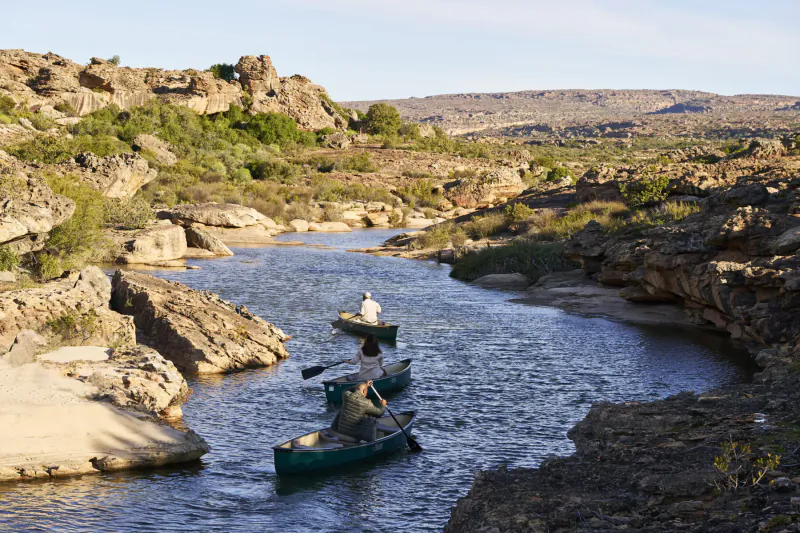 Two green canoes with paddlers navigate a winding river through rocky canyon landscape in Bushman’s Kloof