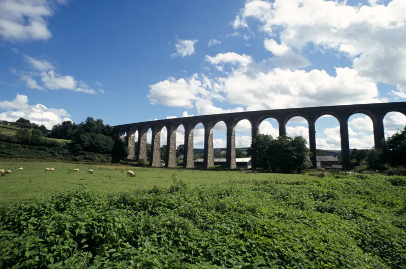 Historic stone railway viaduct with multiple arches over green fields with sheep, blue sky and clouds