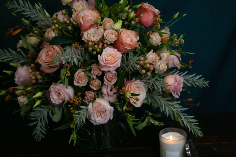 Lavender Green florist bouquet of pink roses, berries, and greenery in vase beside lit white candle on dark table.