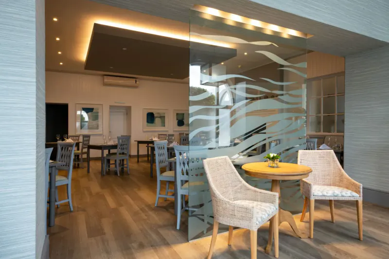Elegant hotel gallery lounge with wicker armchairs around a small table with plant, glass wave partition, and wooden floors.