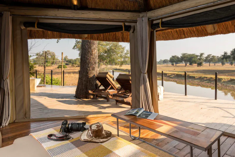 Open-sided thatched Lion Camp deck with lounge chairs overlooking river, savanna, trees; table with camera, hat, book.