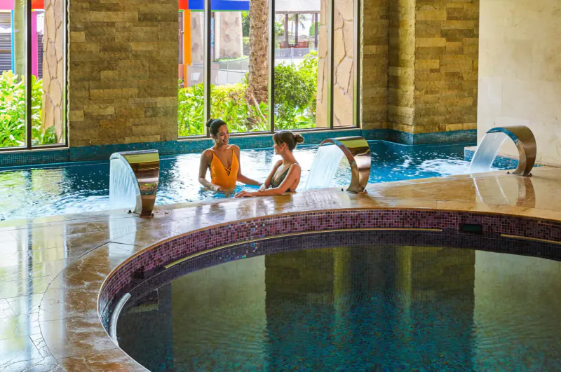 Two women in swimsuits chatting in a luxurious turquoise hot tub at Sofitel Dubai The Palm spa, with stone walls and plants.