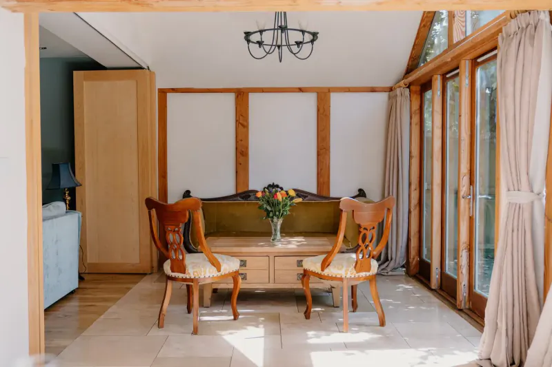Elegant sunroom in Cotswolds country house with carved wood chairs, floral vase on table, chandelier, and large windows.