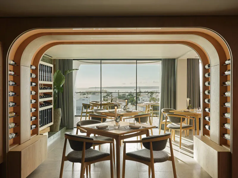 Elegant dining room at W Algarve Hotel with arched wooden frame, wine racks, round tables, chairs, and ocean view.