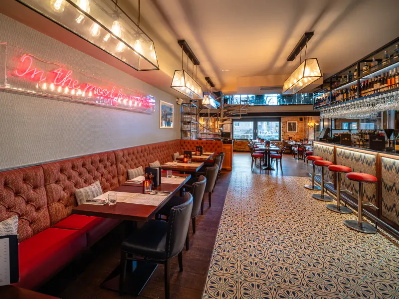 Sophisticated hotel gallery restaurant with 'In America' neon sign, pink banquettes, wooden tables, bar stools, and pendant lights.