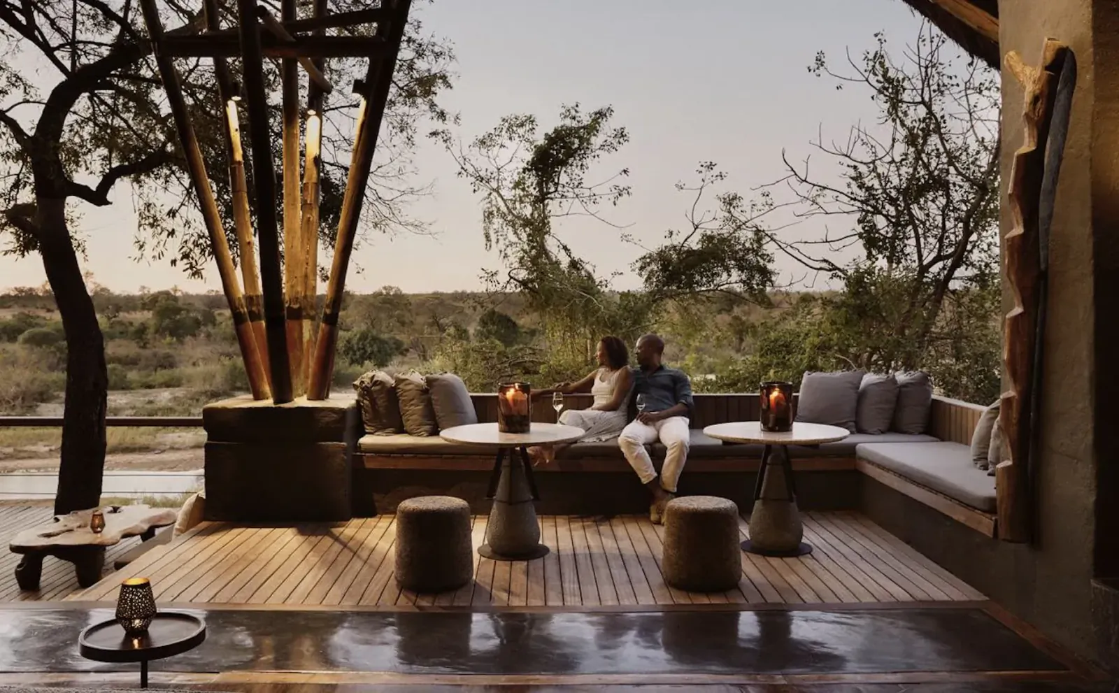 Couple sits intimately on plush outdoor sofa at dusk in African savanna lodge, lanterns lit, bushveld view.