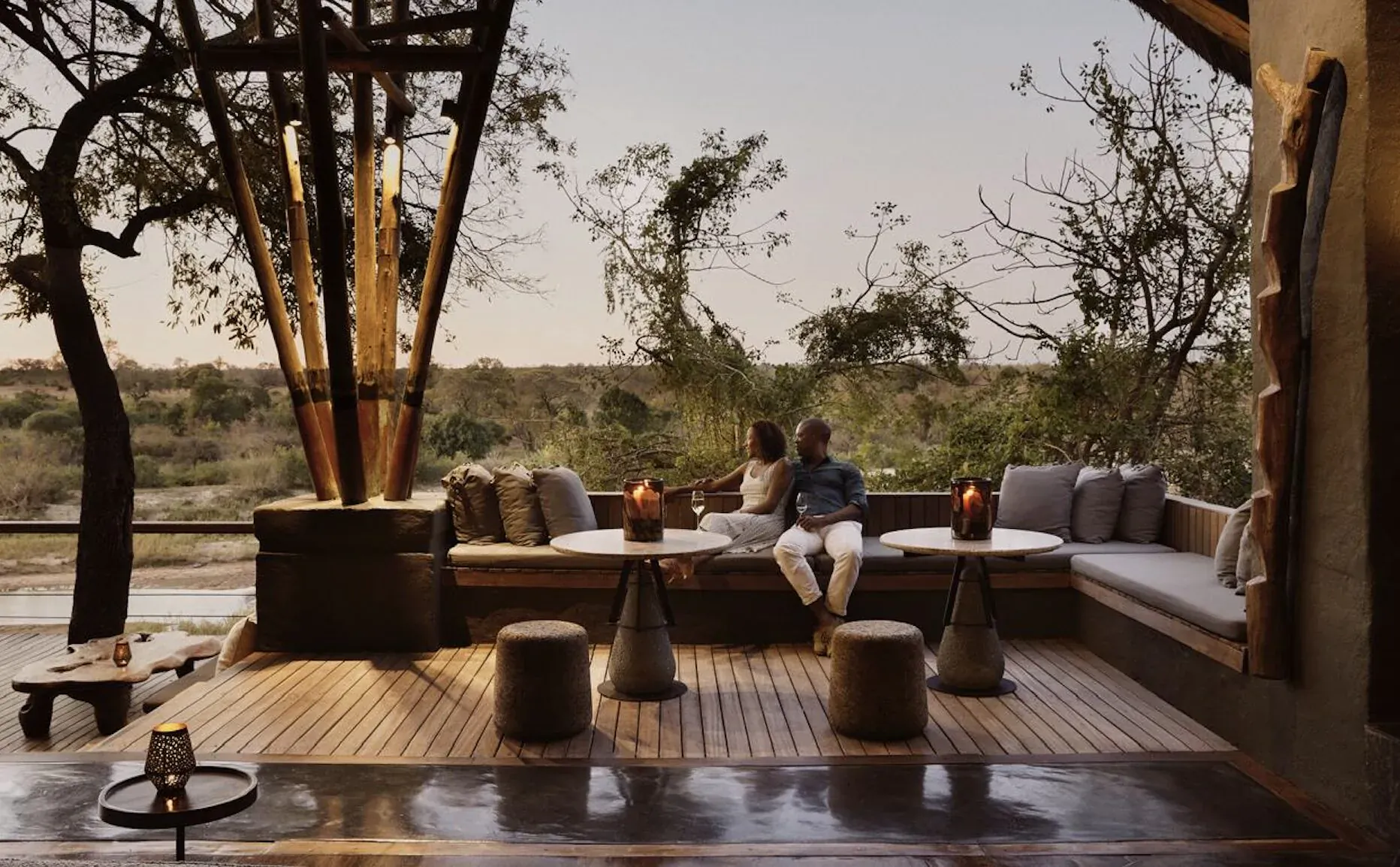 Couple sits intimately on plush outdoor sofa at dusk in African savanna lodge, lanterns lit, bushveld view.