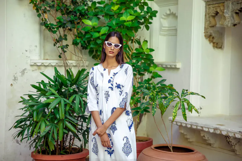 Woman in white kurta with blue paisley prints stands with hands clasped amid potted green plants in arched architectural setting
