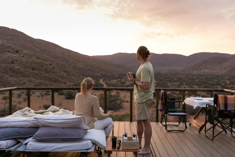Couple on Tarkuni Star Bed deck at sunset, woman seated on bed, man standing holding mug, overlooking arid mountains
