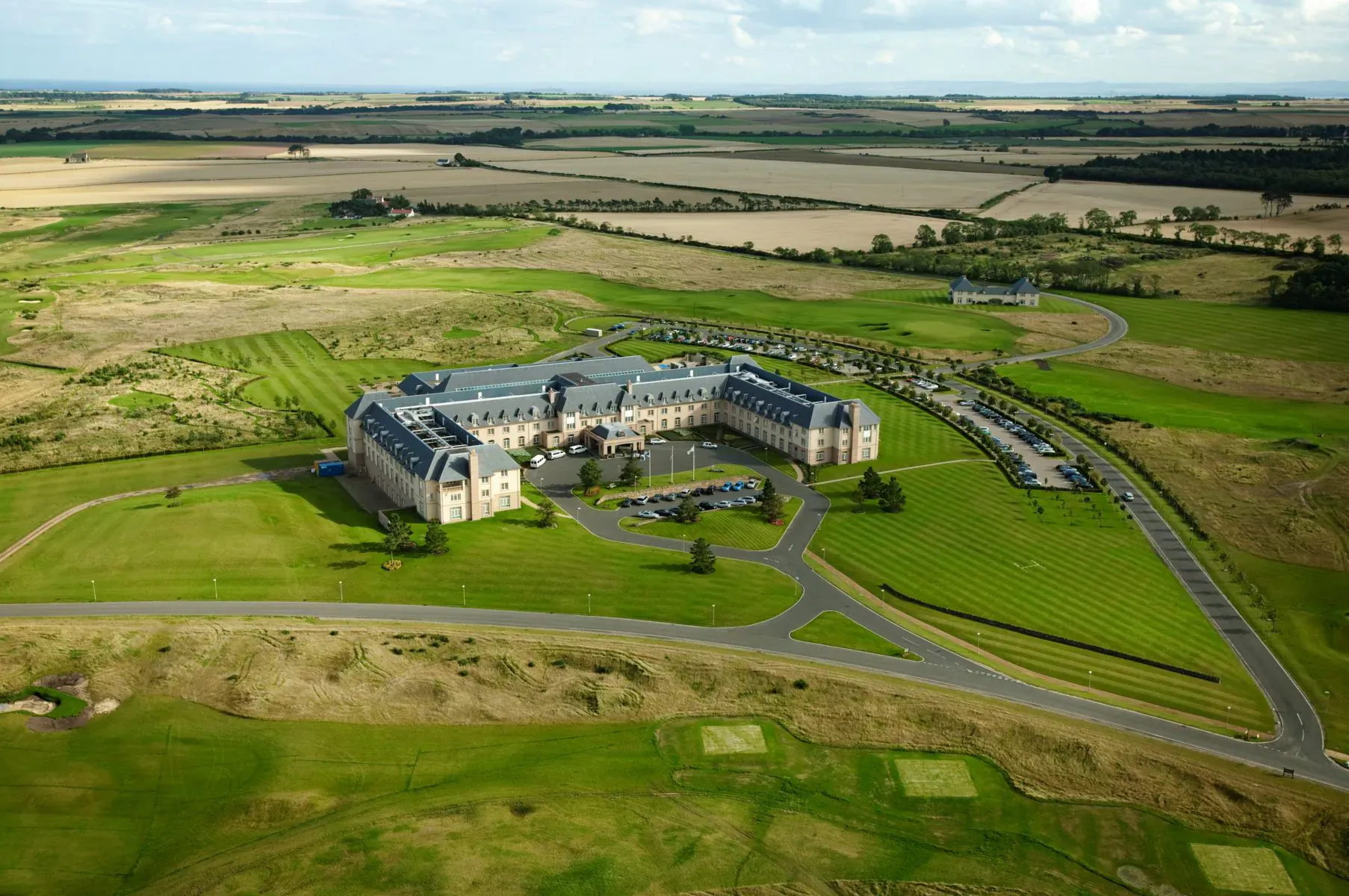 Aerial view of a large château-style building on expansive green golf course amid Scottish fields.