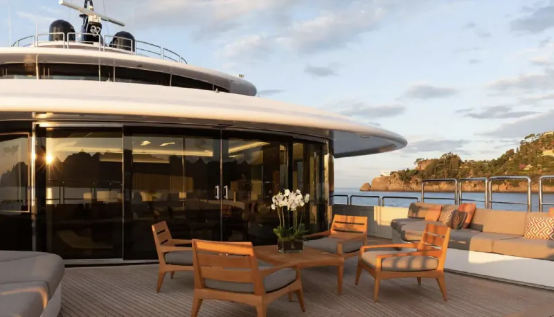 Luxury yacht deck with wooden dining table, chairs, lounge seating, white orchids, glass walls, sea and cliffs beyond.