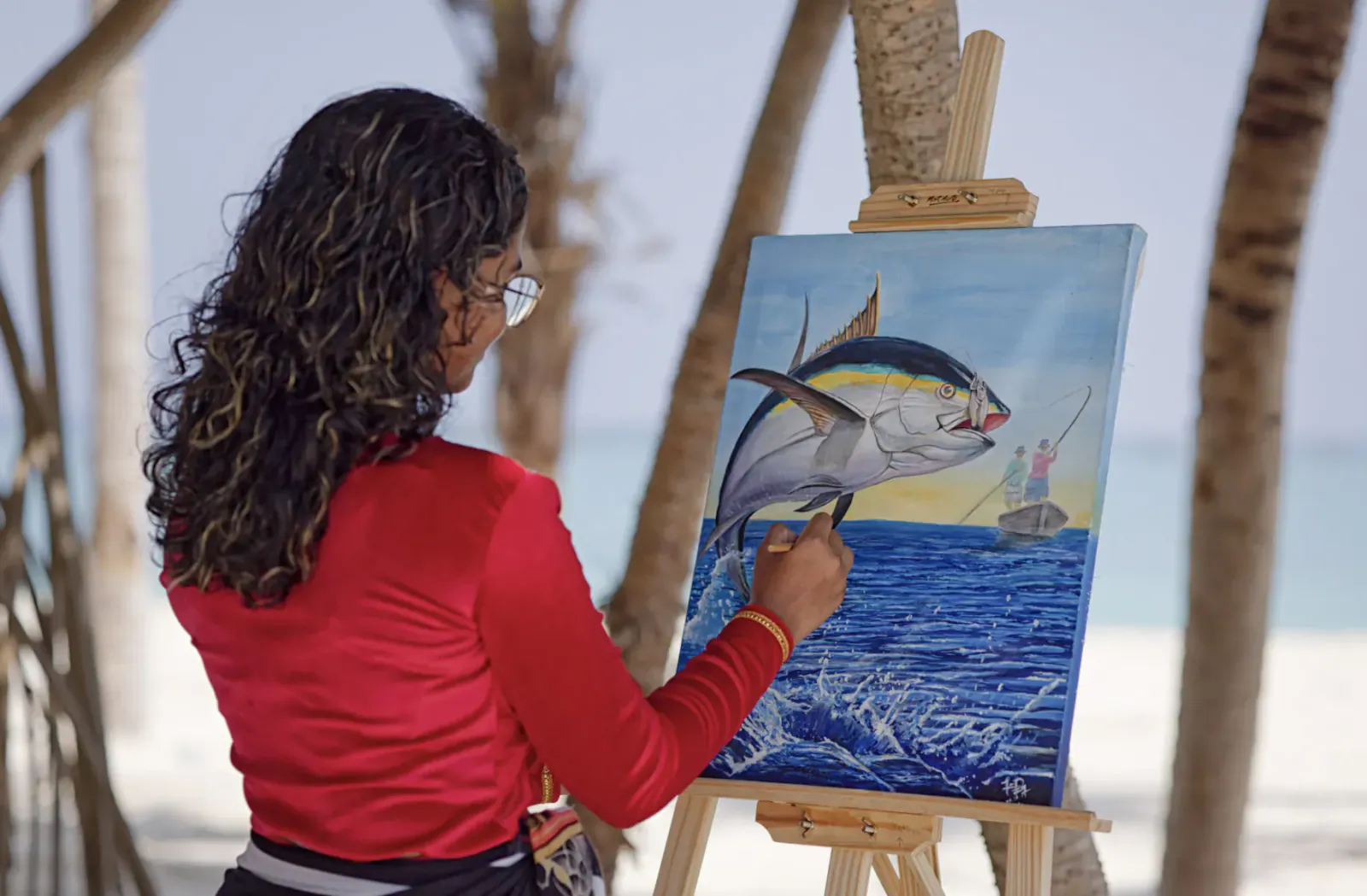 Woman with glasses painting vibrant tuna jumping from ocean on canvas, at Maldives beach under palm trees