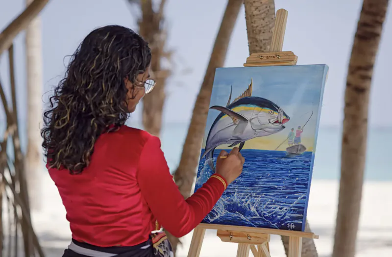 Woman with glasses painting vibrant tuna jumping from ocean on canvas, at Maldives beach under palm trees