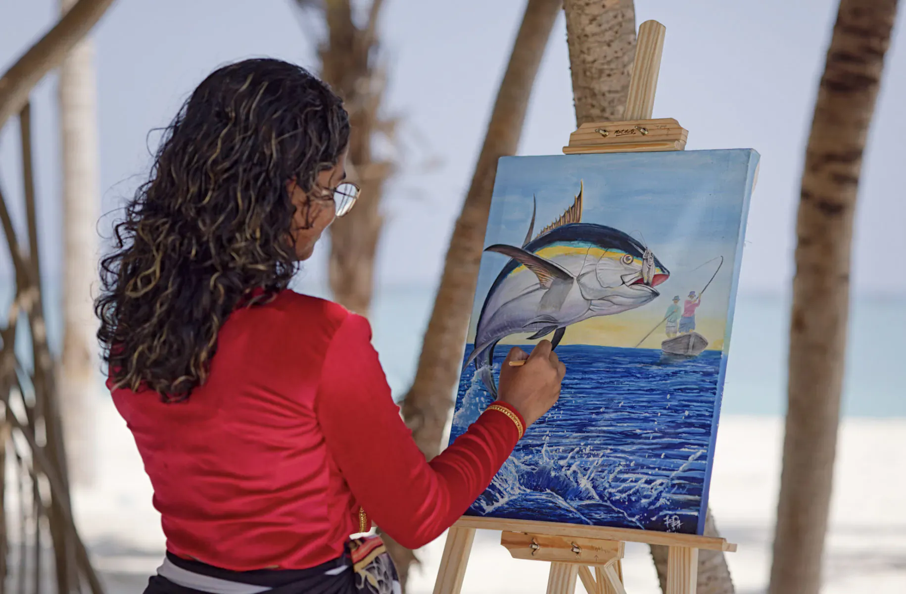 Woman with glasses painting vibrant tuna jumping from ocean on canvas, at Maldives beach under palm trees