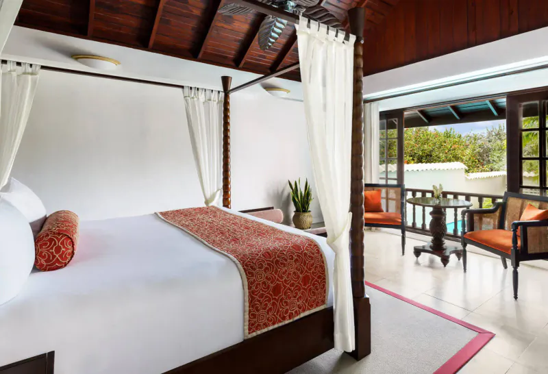 Luxurious four-poster bed with white drapes and red bedding in Spice Island Beach Resort room, balcony view of tropical garden.