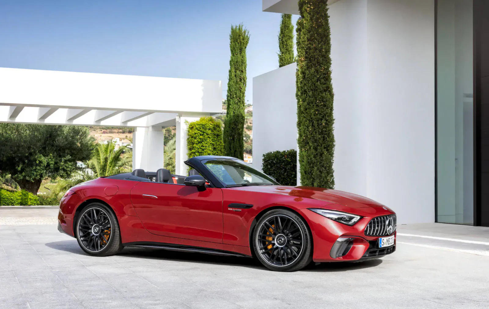 Red Mercedes-AMG SL convertible parked in front of modern white building with cypress trees and landscaping.