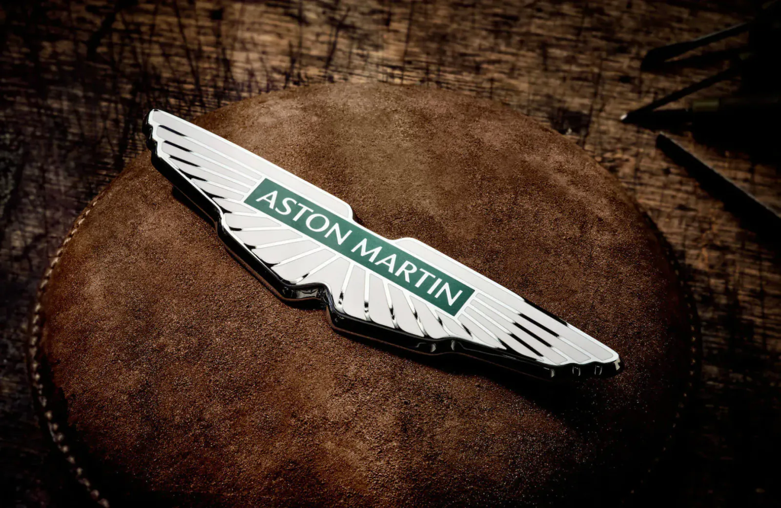 Aston Martin wings logo emblem with 'Aston Martin' text on tan leather over wood background