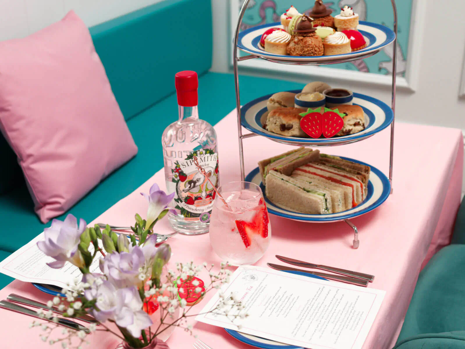 Three-tiered afternoon tea stand with sandwiches, scones, cakes; gin bottle, pink gin, flowers on table.