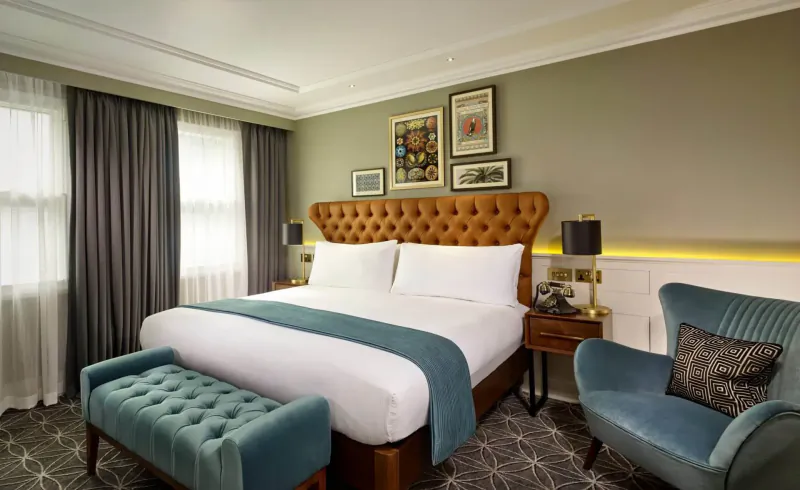 Elegant hotel bedroom with tan leather headboard bed, teal duvet, velvet chair, ottoman, and framed art on walls.
