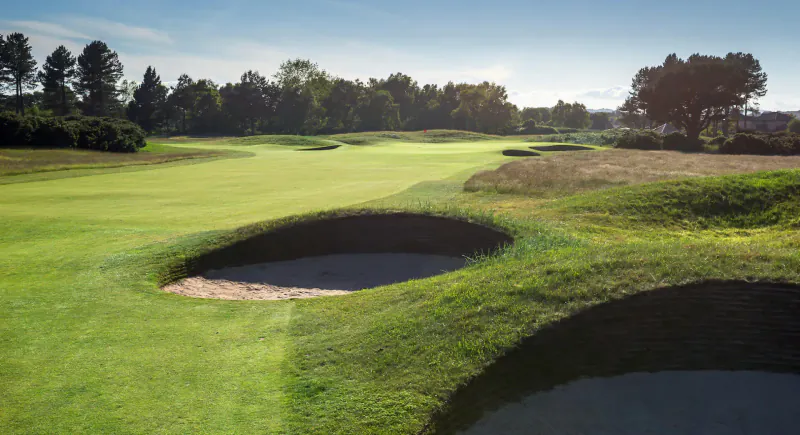 Carnoustie Links golf course with multiple bunkers on lush green fairway, surrounded by trees under blue sky.