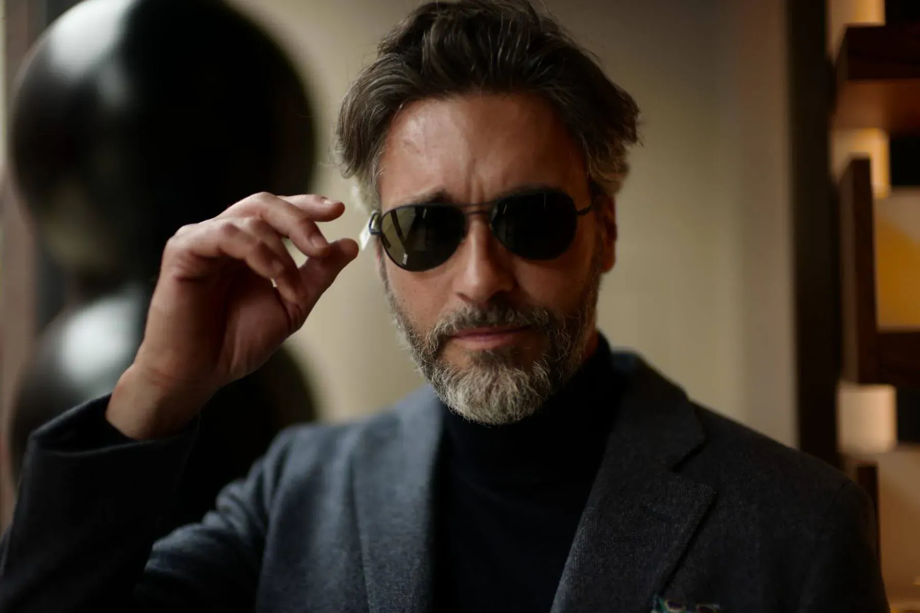 Man with gray hair and beard wearing sunglasses and gray suit, adjusting frames in modern interior