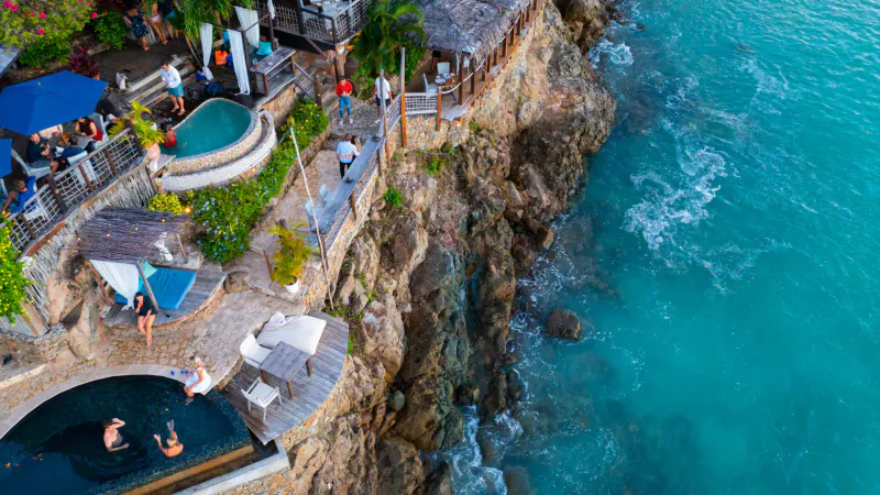 Aerial view of Sheer Rocks resort in Antigua: infinity pool on cliff edge, turquoise ocean, guests relaxing by hot tub and bar.
