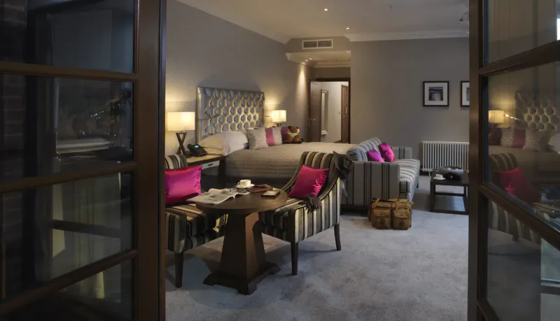 Elegant hotel suite at Northcote Hotel with tufted bed, striped sofa, pink pillows, and open doors