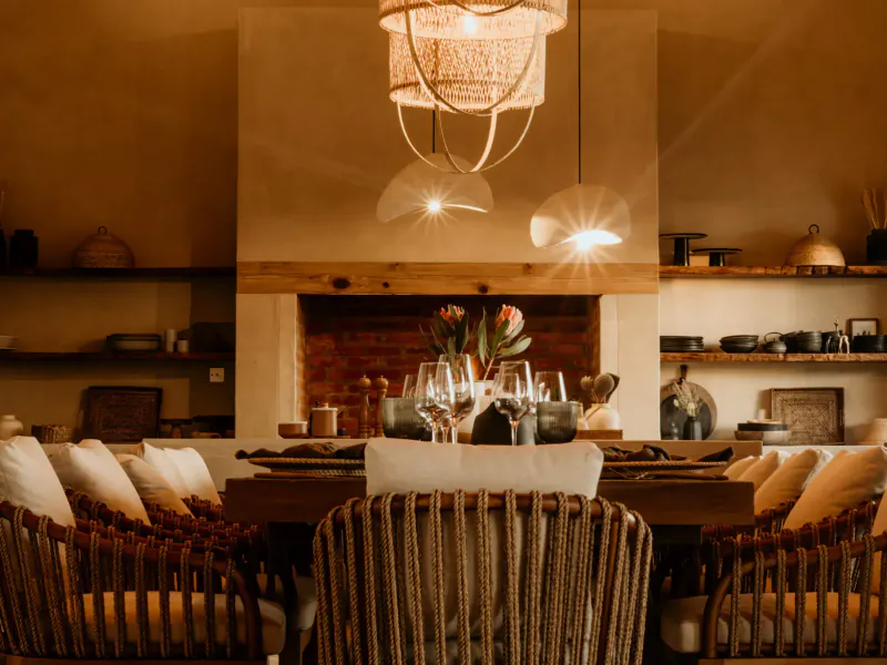 Elegant dining room in Namibian savanna villa with rattan chairs, set table, wine glasses, fireplace, and woven pendant light.